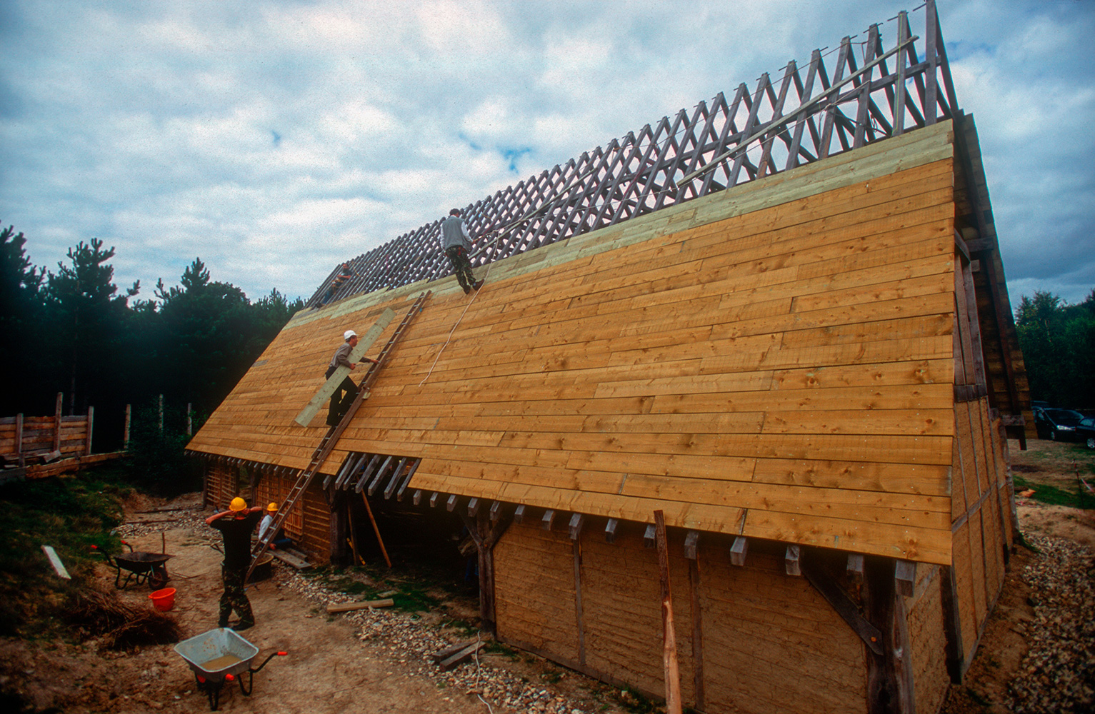 The softwood sarking boards being put on ready for shingling with oak.This is from the Wildwood Trust website;{quote}The site was cleared in 2001 and work on the Longhall commenced at Easter the following year. At the end of 2004, the main structure of this 20m × 10m × 10m building was completed and before the onset of winter, we had clad most of the roof with softwood sarking boards, awaiting the first 10,000 hand-cleft oak shingles for delivery in 2005. That year saw the completion of the porches and the commencement of the huge task of nailing on the shingles. Walling advanced well, too. In 2006, the front roof was completed and shingling advanced about a third of the way up the back roof. The walls were completely infilled, doors and hinges were fitted and our hall was secure for the first time. 2007 saw the completion of shingling, some 18,000 eventually finding their way onto the roof.It is an impressive sight and is certainly the largest reconstructed early medieval building in private hands in Europe. Built entirely of English oak, all of which has been harvested from trees growing in Kent, (many within a couple of miles of the site), the site in general and the Longhall in particular, has been conceived, planned, purchased, designed and constructed by our members. What you see is the result of many long hours of research, discussion and the practical application of skills with which our ancestors would have felt at home.{quote}