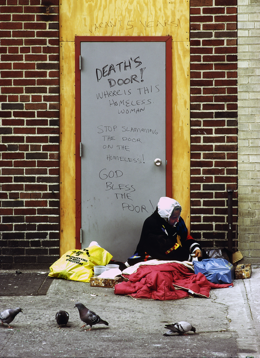May 1988 - Homeless woman living on the streets of New York City in front of a door covered with graffiti condemning homelessness.