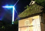 A grass roofed hut is juxtaposed with a modern wind generator that is illuminated with a blue spotlight.