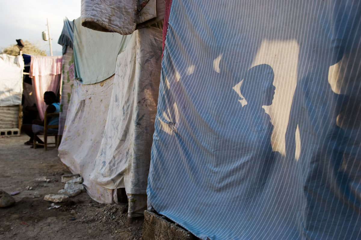 Five year-old Jonas Joseph, his eight year-old sister Marie and 12 year-old brother Jeff are silhouetted against the wall of their family's makeshift shelter, in the midst of a tent city that serves as a temporary home for 350 families.  The earthquake caused heavy damage to residential buildings in Carrefour; an estimated 80-90% of the buildings were destroyed.  © Habitat for Humanity International/Ezra Millstein