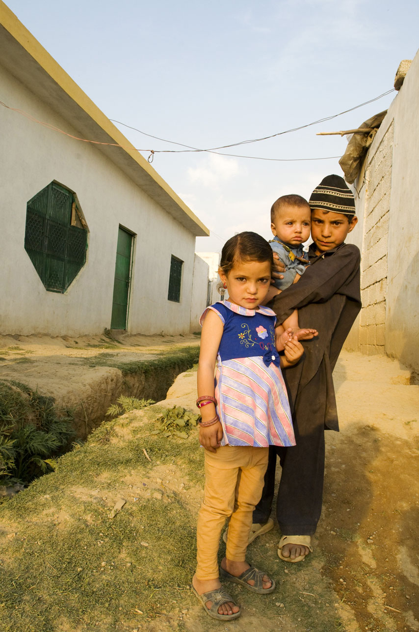 Jafir (far right), 7yrs old, holding his baby brother Kamran, and standing next to his sister Asma (left), 4yrs, inside a refugee community set up on the edge of Islamabad, Pakistan.  These children and their parents fled their home  in Bajaur 4 months ago and have no idea when they will be able to return. This refugee community that they are living in now was set up by refugee, Bir Baba, also from Bajaur, who was forced to leave his home when the Taliban threated to kill him if he did not leave his village.  There are now 70 -80 undocumented refugee families living in the community.