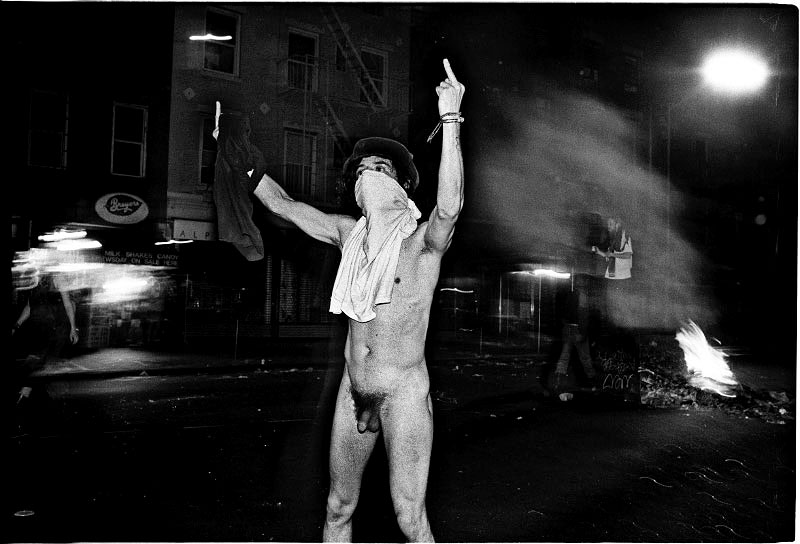During a protest in the Tompkins Square Park, a protester indicates double insulting signs at NYPD’s riot geared forces. His being naked is also an intentional protest, since NYPD, then, is recognized very brutal by many residents of the community. New York, May 27, 1991. 