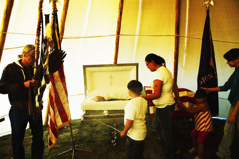 A scene of the funeral service for Vern American Horse, 83 year old Lakota who died due to diabetes. Diabetes is one of the biggest problems in Native American reservations like in Pine Ridge, since the residents have nearly no-choice, unhealthy dietary habits, plus due to the possible radioactive water contamination. (Vern American Horse was a resident in Pine Ridge, but the funeral was held a nearby town of Gordon, Nebraska.) Plus, due to the fact that the practice of native American religious ceremonies had been banned in the late 19 century to the 1970s, many Lakota people are Christians, though now the youths have started to have their own traditional faith.