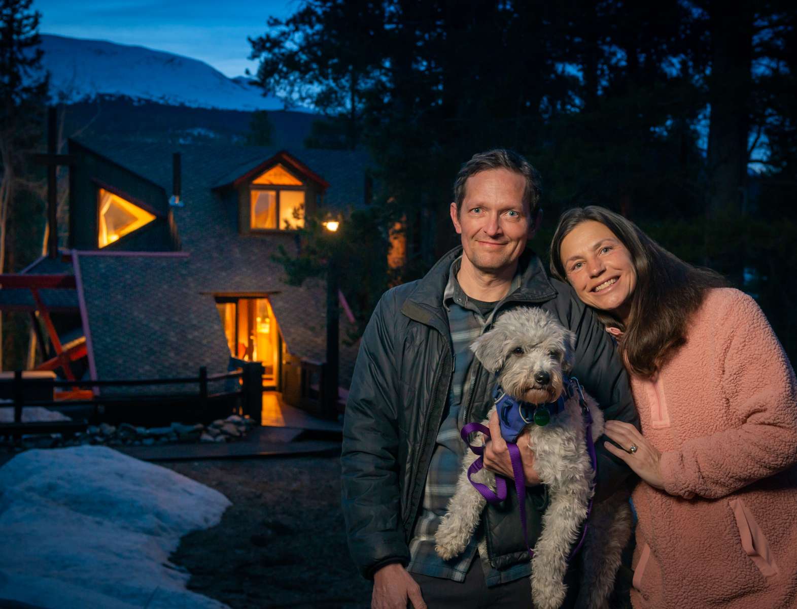 A Colorado couple with their dog and best friend, Sunny, at dusk in front of their unique mountain cabin  as winter gives way to mud season.