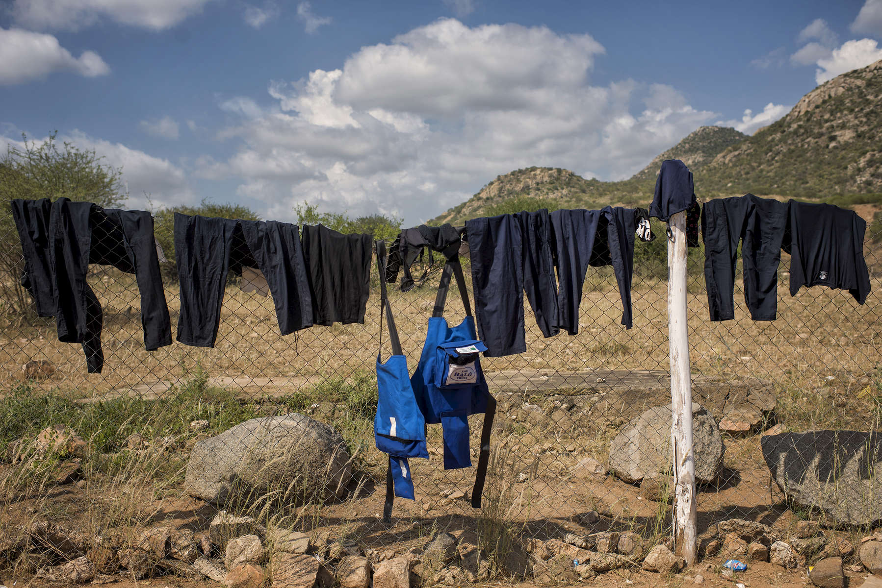 May 5, 2018: KANENGUERERE, ANGOLA - Clean HALO uniforms and body armor hang from the perimeter fence to dry at the end of the working week.  These deminers are working under extremely difficult circumstances in Kanenguerere.  Not only is extremely hot - with snakes and scorpions common - but much of the mined area is on the side of an extremely steep hill, making every step dangerous. The area was mined during the civil war by government forces to protect the nearby railway line that can be seen in the background, as well as various troop positions. It is currently used by roughly 170 people including village residents and nomadic herders - many of whom are young children - who pass through uncleared land every day. 