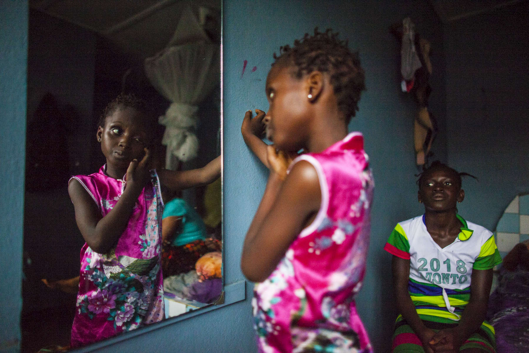 Ebola survivors Aminata Conteh, 8, looks in the mirror as Isatu Tholley, 12 looks on at their guesthouse in Freetown, Sierra Leone.