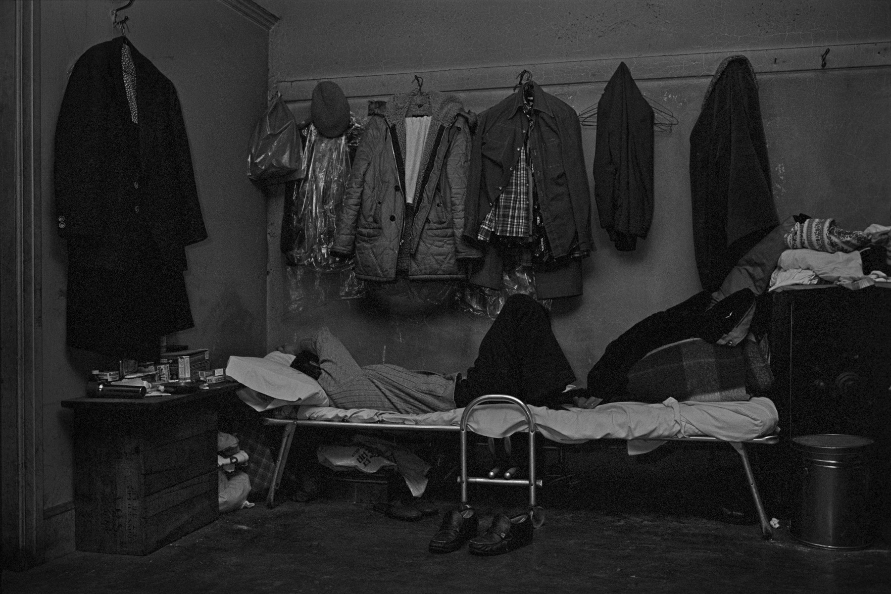 A man sleeps on his bed in the Bachelor Apartment he shares with 3 other men, Bayard St., New York Chinatown, 1982. He is surrounded by his belongings, including coats and jackets hanging on the wall above him.