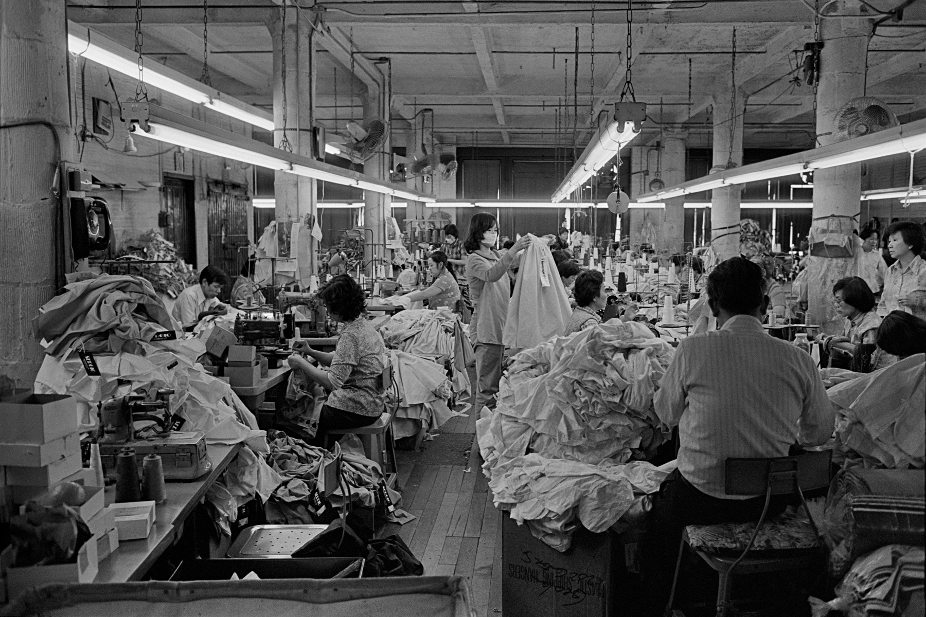 A view of Geolan Sportswear a Garment factory, 202 Centre St. New York Chinatown, 1983. Numerous people are shown working in the factory that is lit with flourescent lights. 