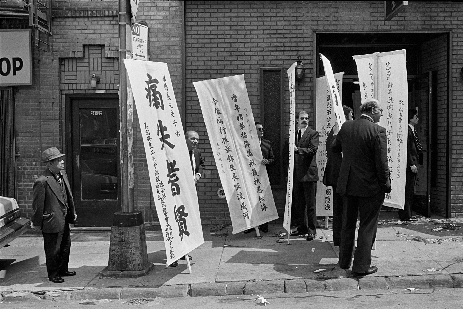 Funeral, Mulberry St., New York Chinatown, 1982.The part of Mulberry Street next to Columbus Park used to be home to funeral homes run by Italian Americans when Little Italy stretched south of Canal Street. After Chinese moved into the area and took over the funeral businesses, some of the Italian workers stayed on. This photograph shows a funeral with Chinese banners being held up by non-Chinese funeral home workers.
