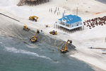 Heavy equipment is brought in to fortify the shallow shoreline and threatened property against rising seas and extreme weather.  A pipe (upper left) feeds in sand that is pumped from the bay to the equipment building up an artificial sand barrier.