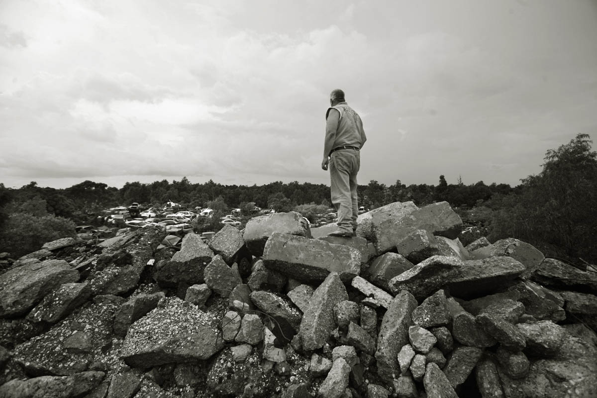 Roger Cull , of Stonehedge Construction a cement recycling business,, was vacationing in New Orleans with his family when Katrina threatened the city. Two days after she hit, he brought his construction company to New Orleans to begin the clean-up. He stands on chunks of New Orleans foundations, cement from houses, roads, and foundations destroyed by Katrina. His company has 350,000- 400-000 tons of recycled cement in piles, and from the top of one; a vista of his neighbor's junk yard where Katrina cars are being stored until the price of metal goes up in the future.