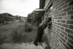 A portrait of community leader Aaron Broussard, resident and member of the East Shore neighborhood association concerned for his neighborhood and the blight he is seeing from absent residents and overgrown properties. He looks through a window of an empty house that still has debris from Katrina inside. 