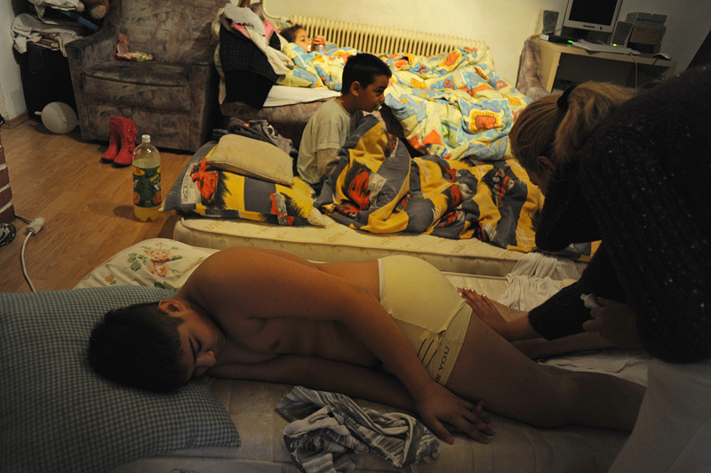 DEVECSER, HUNGARY.  (Far right) Ilona Autal, 40, rubs cream on the legs of her son, (left) Peter Bogdan, 12, as her other children, (on sofa) Miriam Melitta Bogdan, 3, and (center) Renato Achilles Bogdan, 5, wake up and watch television  before school in their living room on November 25, 2010.  Peter was walking home when an industrial reservoir containing toxic red alumina sludge broke at the MAL plant in nearby Ajka, Hungary and sent a torrent of the hazardous material gushing through the streets of Devecser and consequently has burns on his legs.