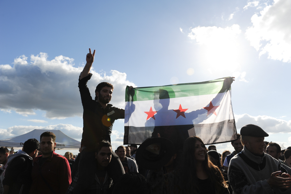BERLIN, GERMANY.  Syrian refugees and others dance and sing and hoist up the Syrian flag at a {quote}Refugees Welcome{quote} picnic for refugees and asylum applicants arriving in large numbers in their country after having fled conflicts in the Middle East, Afghanistan and elsewhere at the Tempelhof Airport Park on September 27, 2015.  German Chancellor Angela Merkel has said the country is prepared to accept a historic 850,000 asylum applicants this year but has also sought to tighten its border control with Austria and other Schengen zone countries.