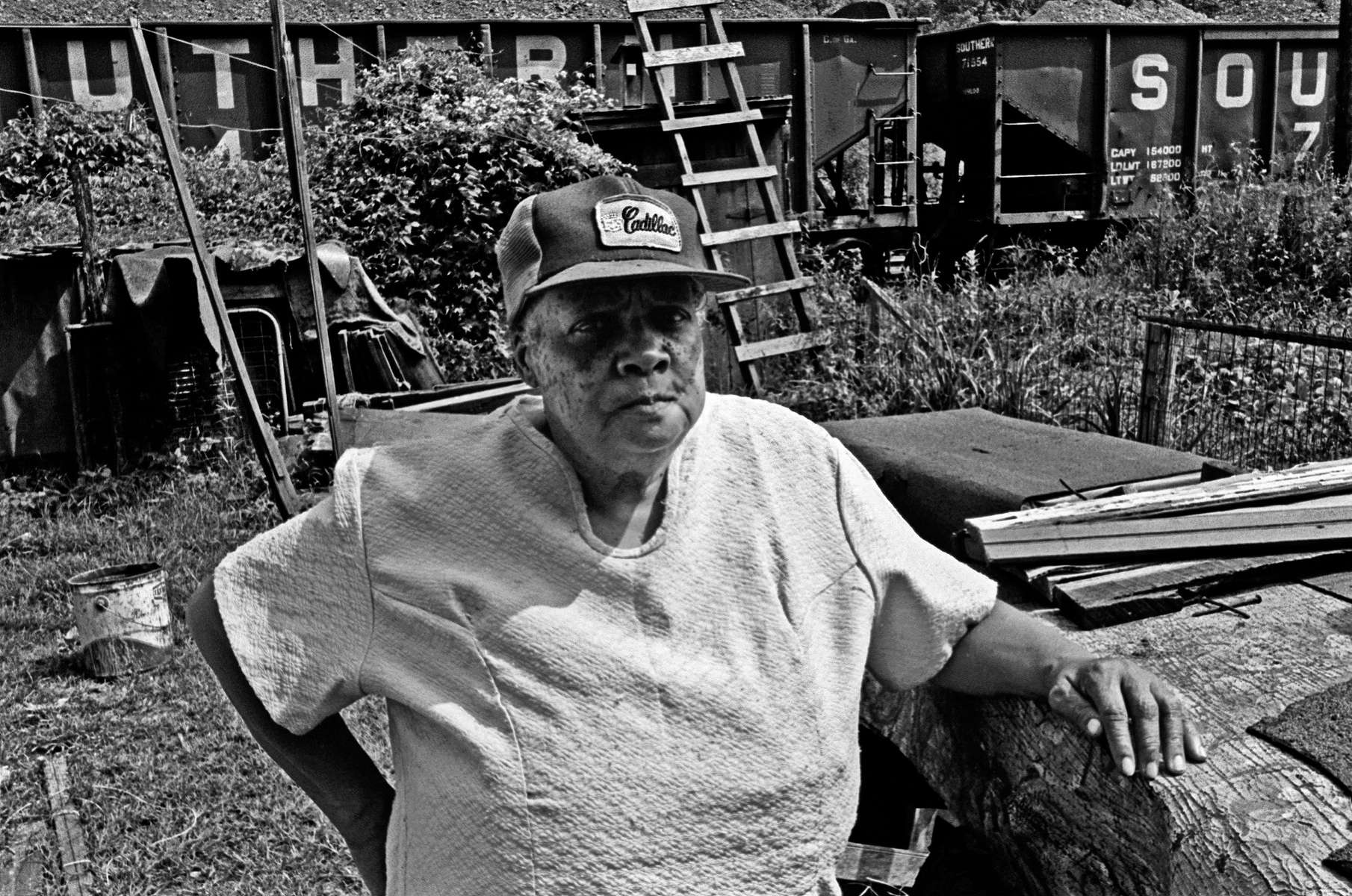 Woman with a Cadillac hat stands in her backyard, with a freight train parked on tracks behind. She lived in an all-Black hollow in the back hills of West Virginia. She posed willingly for me, but after few minutes her husband called her back into the house. She quickly told me, {quote}You'd better get moving. My husband says he's getting his gun right now.{quote} People don't take kindly to strangers, and no stranger has any reason to visit this small, isolated settlement, unless they are a federal agent or a drug dealer. Jon Chase photo 