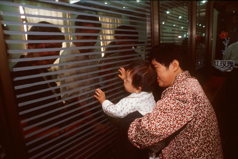 Adoptive parents wait behind a window to receive their child, in the arms of an adoption worker.
