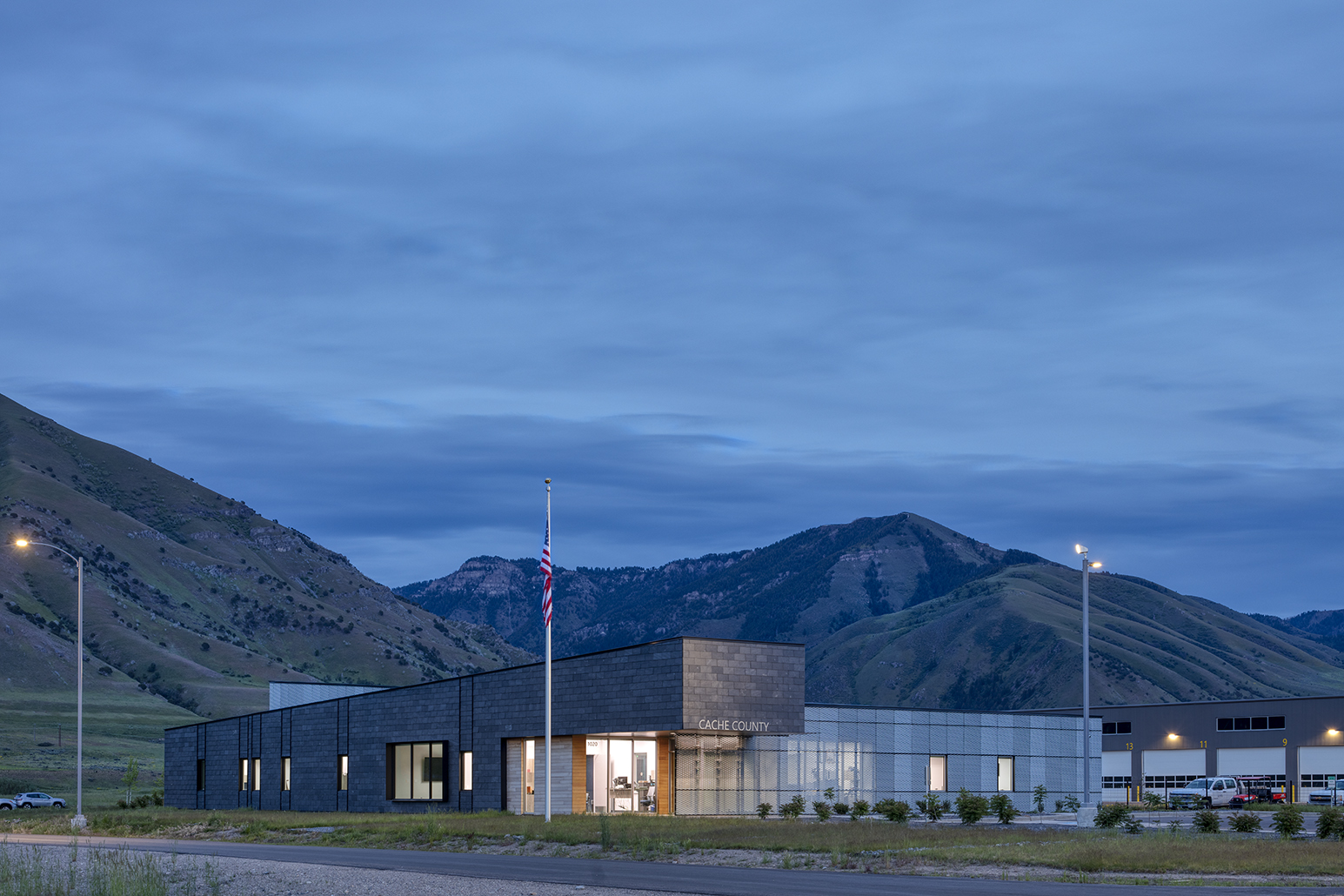 Exterior view of the Cache County Public Works Facility nestled up against the mountains in Hyrum, UT. The building is a metal and slate clad structure that is low in profile and modern looking and the picture was captured at dusk with the interior lights on.