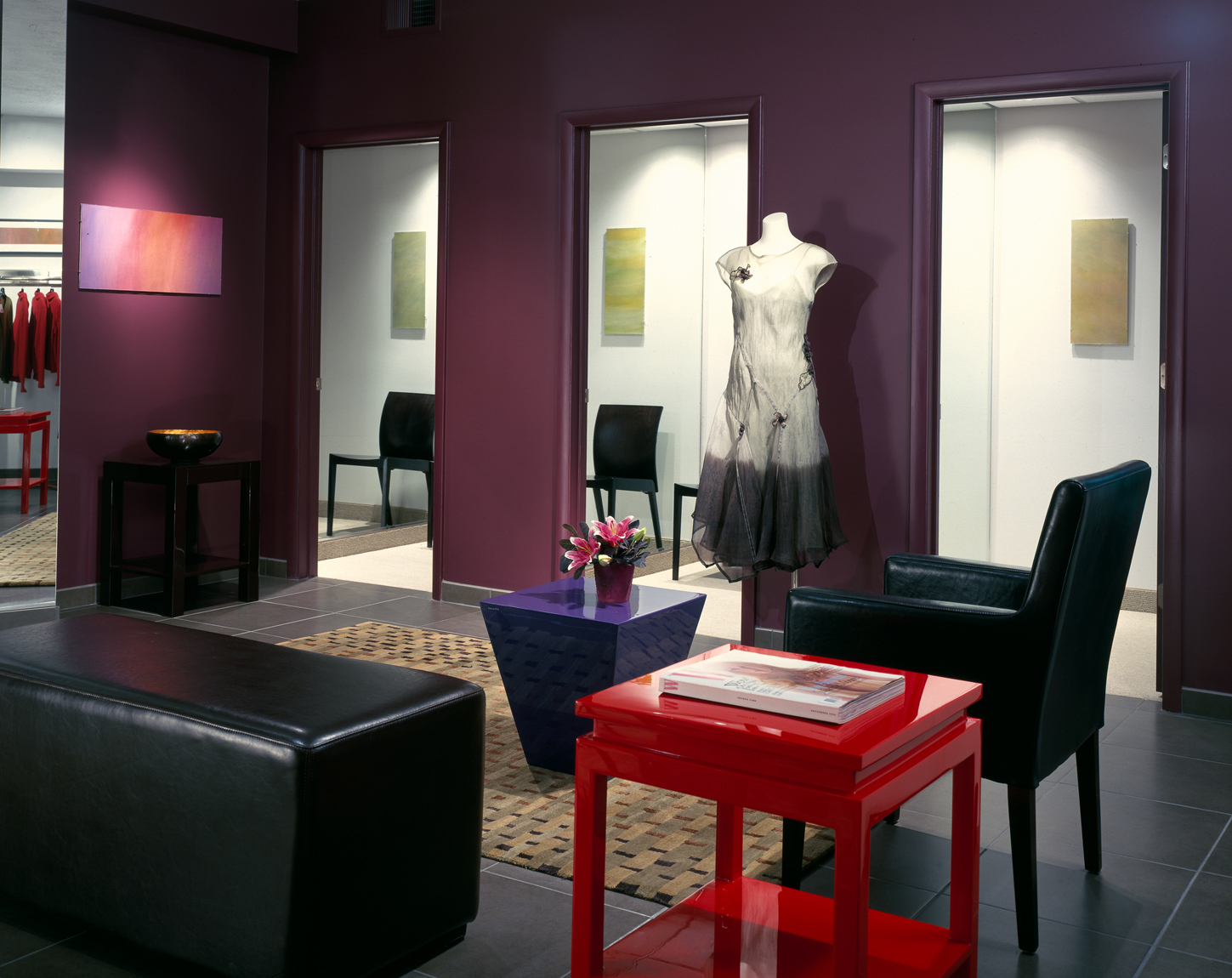 Interior view towards changing rooms in an upscale clothing boutique. Black leather furnishings are framed by maroon walls and the centerpiece features a bright red table.