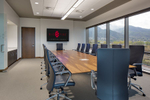 This photograph shows an interior view of a corporate boardroom with a long wood boardroom table, a monitor at the end of the table and floor to ceiling glass windows with views of mountains. 