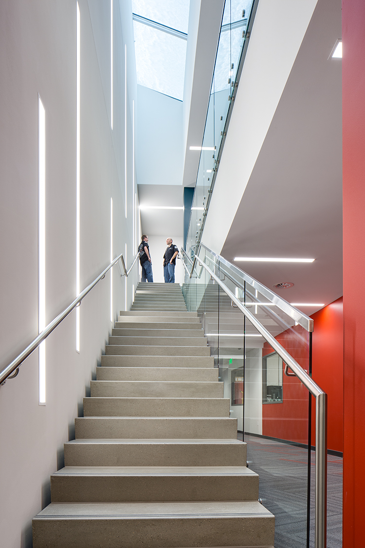 Two people conversing at the top of of long stairwell. The ceiling above the stairwell has a large skylight and the left hand wall has vertical strip lights built in. 