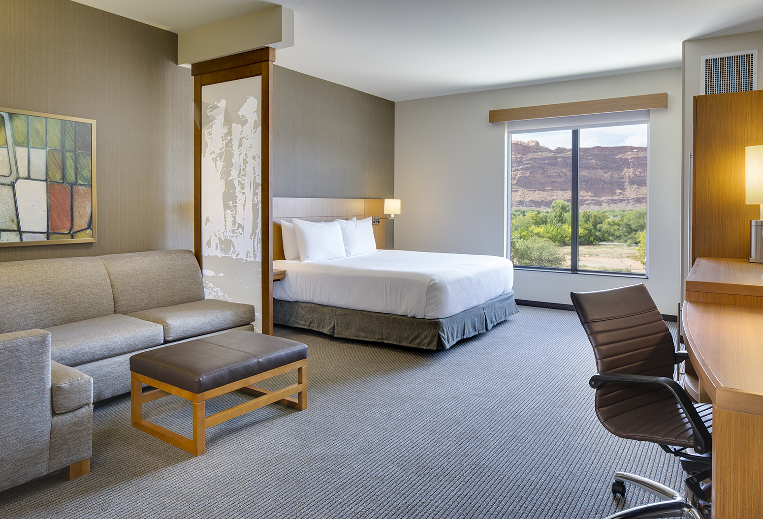 Daylit photograph of a comfortable suite at the Hyatt Hotel in Moab, UT. The viewer sees a modern desk and sofa in the forground with a kingsize bed next to the window that has views of thr red rock country and green cottonwood trees