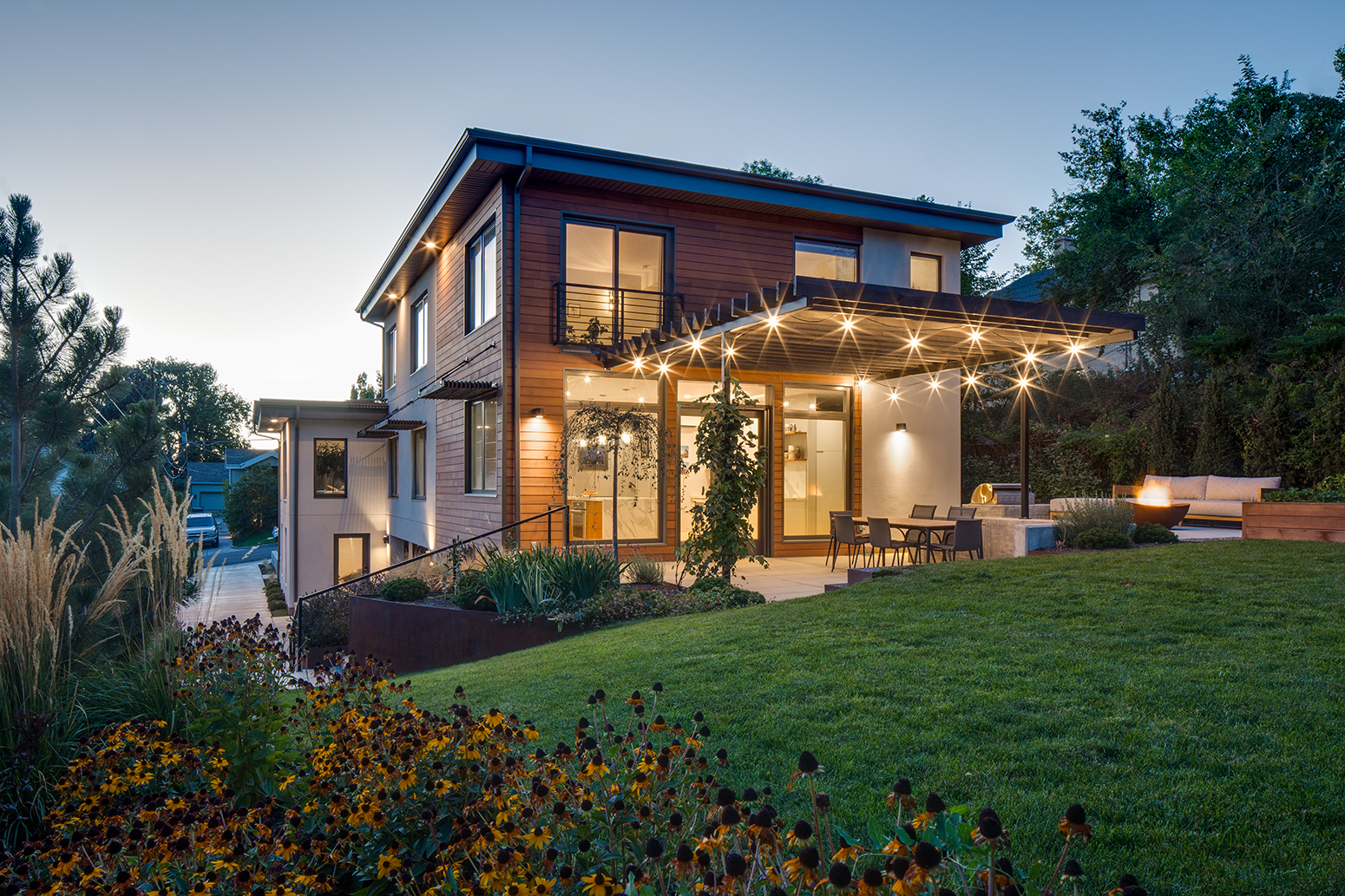 Exterior photograph of the back veranda and yard of a modern home in a residential neighborhood known as the Avenies at dusk in Salt Lake City, UT. Ther veranda has sparkling lights draped above the concrete sitting area with an open fire pit lit and flames. 