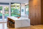 This is a photograph of a kitchen in a modern home with a large marble island with a bowl of red timatos on it.. Through the windows and doors the viewer sees the spacious, green back yard.