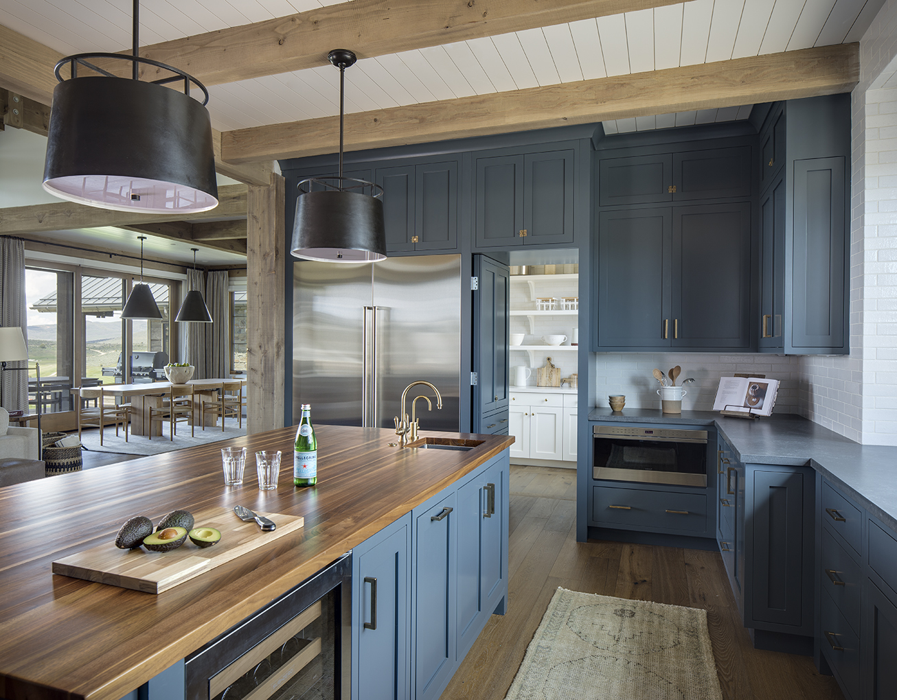Interior photograph of a residential kitchen with views beyond to the dining area and beyond to the mountains of Park City, UT. All of the cabinets are blue and the ceiling is white bead board with exposed timber beams.