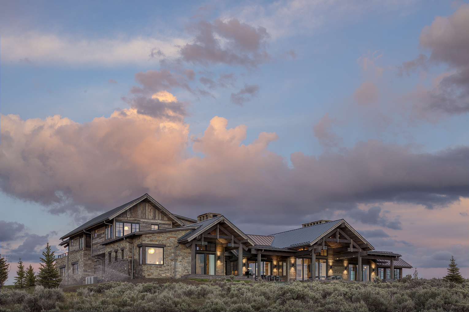 Exterior photograph of a private residence near Park City, UT. The house is surrounded by sage plants and dratic skies at dusk and the home is clad in reclaimed wood that looks very natural.