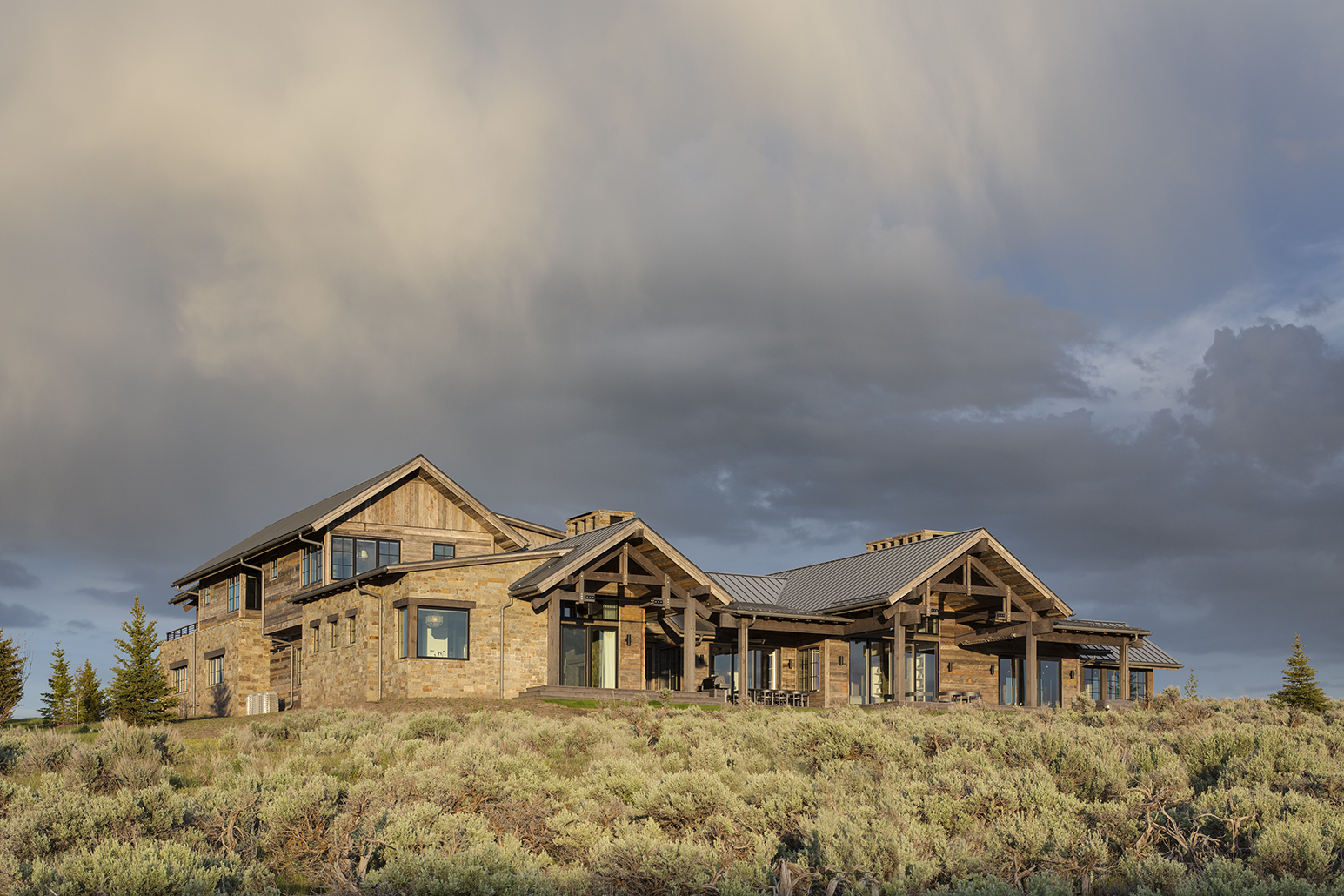 Exterior photograph of mountain rustic luxury residence at sunset near Park City, Utah. The photograph shows the main facade of recalimed timber, bathed in warm afternoon light, with dramatic and stormy skies behind the building. The home is surrounded by sage.