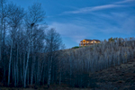 This is a long distance photograph of a private residence of a rustic home positioned on a ridgeline at dawn. The home radiates a warm orange glow and is framed by trees that have lost their leaves due to the time of year