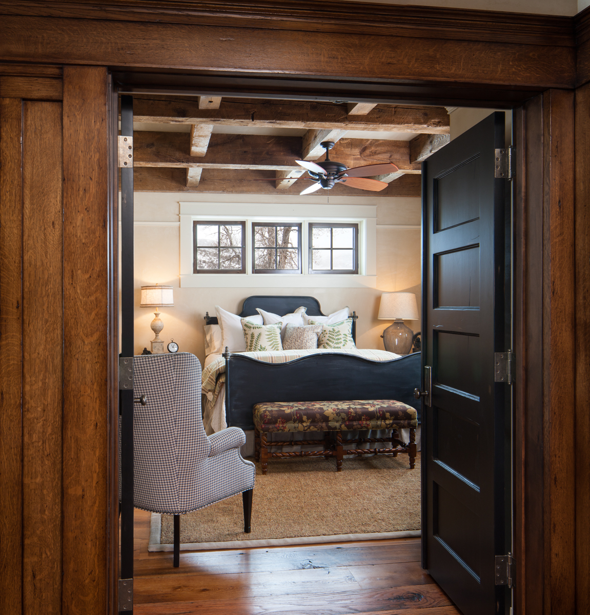 This is a photograph of a view into the master bedroom in a new but very rustic mountain home near Park City, UT. There is oak wood pannelling on the outside of the dor leading in and beautiful timbers that cris croos the ceiling above the master bed.