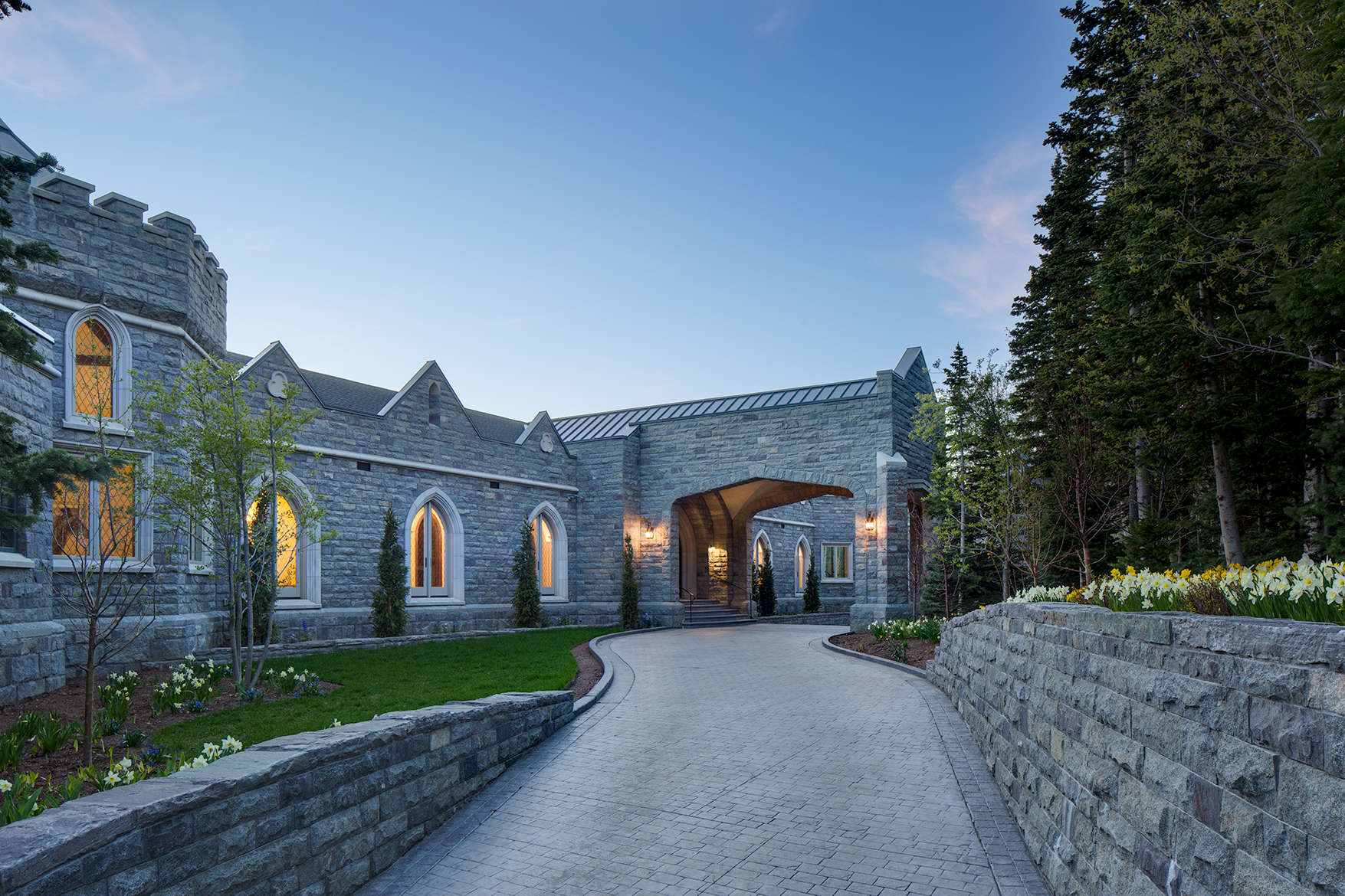 Exterior view at dusk looking up stone walled drive way of a Scottish manor house in Park City, UT with glowing windows.