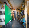 This is a photograph of a man working on his outdoor toys in his man cave at his workbench. The camera is looking down the narrow, room and on one side of the room all of the back pack are hung and on the other side of the room is his work bemch and strorage cabinets with green doors. There are also a few rows of cross country ski boots on the wall facing the camera