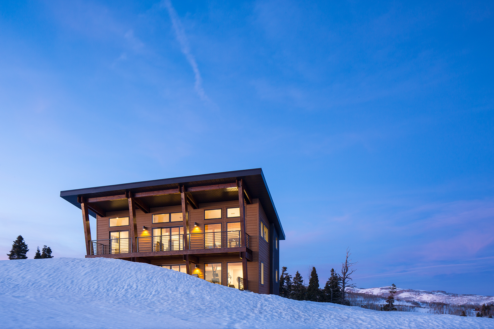 Modern home set high on top of a mountain surrounded by trees and a snow covered meadow. The photograph was taken at dusk so the clouds are oink, the skies are blue and a warm glow is radiating from the windows.