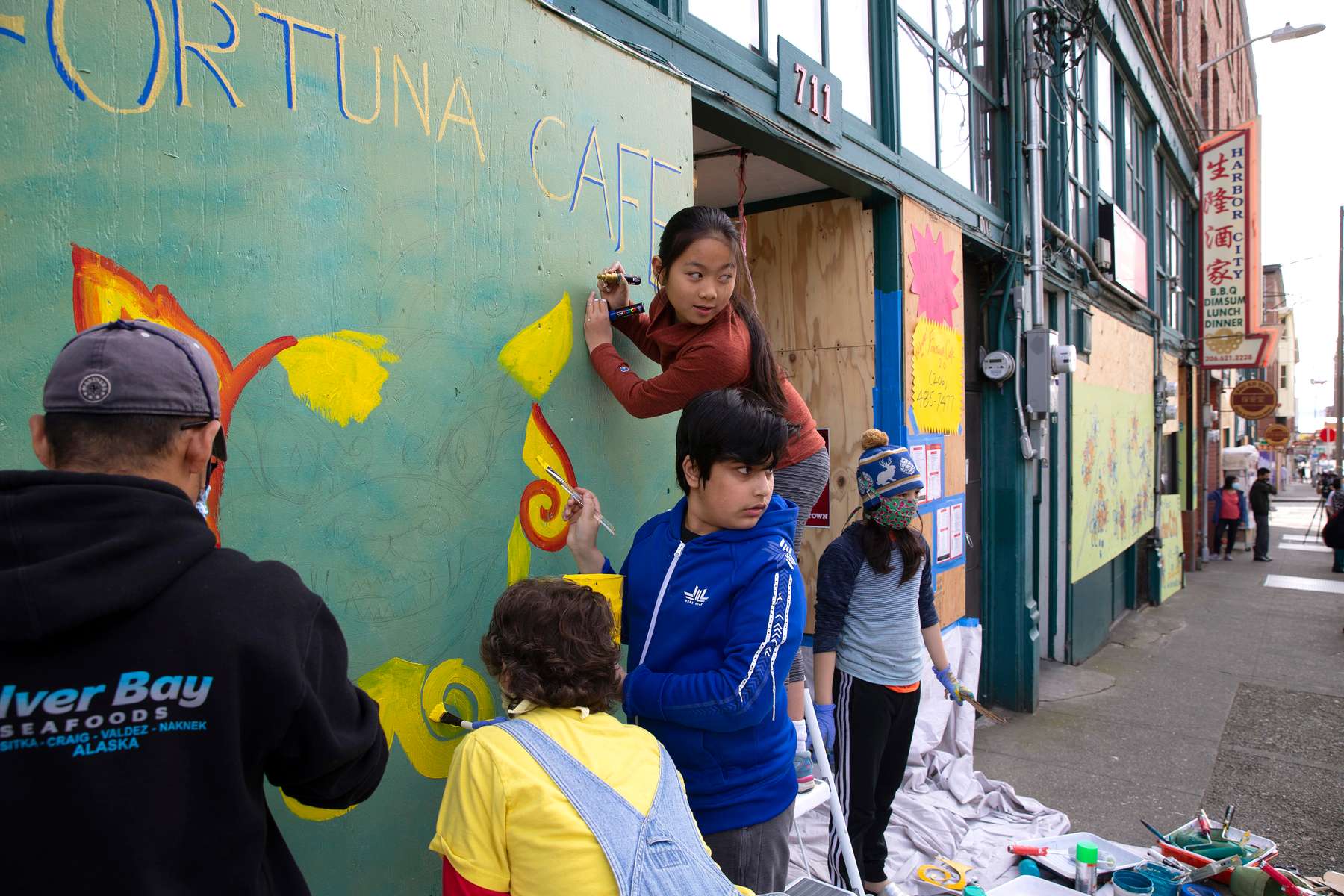 5th graders from Mr. Daichi Hirataís class at the Waldorf School, including Kabir Sethi (center), 11, and Mika Kodama- Chew (on ladder) paint a mural of a Chinese lion over the boarded up storefront of the Fortuna Cafe in Seattle, WA on June 9, 2020. Many storefronts were damaged after a night of rioting swept through the CID after the death of George Floyd, a black man who died when a police officer pinned him to the ground with a knee on his neck. The community responded by boarding up storefronts and hosting artist events to brighten up the streets with murals painted on them. The kids were distracted by firemen putting out a fire at the building across the street. (photo by Karen Ducey)