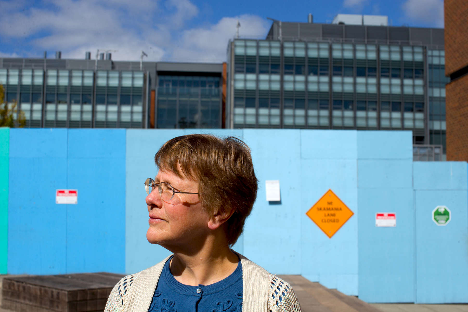 Kathy Bentson, a former research scientist in the Primate Center at the University of Washington, stands in front of a tall, blue wooden wall bearing No Trespassing signs that blocks the public's view of the construction of the new underground animal research lab in Seattle, Wash. on September 21, 2016. In the rear is the William H. Foege Building for Bioengineering and Genome Sciences. Bentson is trying to submit the results of a study she did with others on how to reduce the prevalence of abnormal behaviors in laboratory monkeys to peer review journals, but has met a lot of resistance from the university. (© Karen Ducey Photography)
