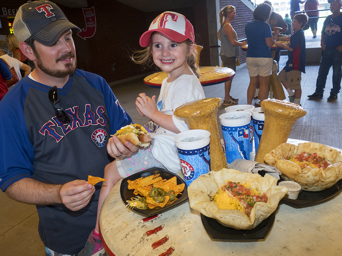 Texas Rangers fans get stuck into some snacks at Globe Life Park ahead of a gamed versus the Los Angeles Angels.Globe Life Park is a stadium in Arlington, Texas, located between Dallas and Fort Worth. It is home to the American League's Texas Rangers baseball team, and the Texas Rangers Baseball Hall of Fame.Dallas is a major city in Texas and is the largest urban center of the fourth most populous metropolitan area in the United States. The city ranks ninth in the U.S. and third in Texas after Houston and San Antonio. The city's prominence arose from its historical importance as a center for the oil and cotton industries, and its position along numerous railroad lines.For two weeks in the summer of 2015, photographer Peter Dench visited Dallas to document the metroplex in his epic reportage, DENCH DOES DALLAS.Photographed using an Olympus E-M5 Mark II©Peter Dench/Getty Images Reportage