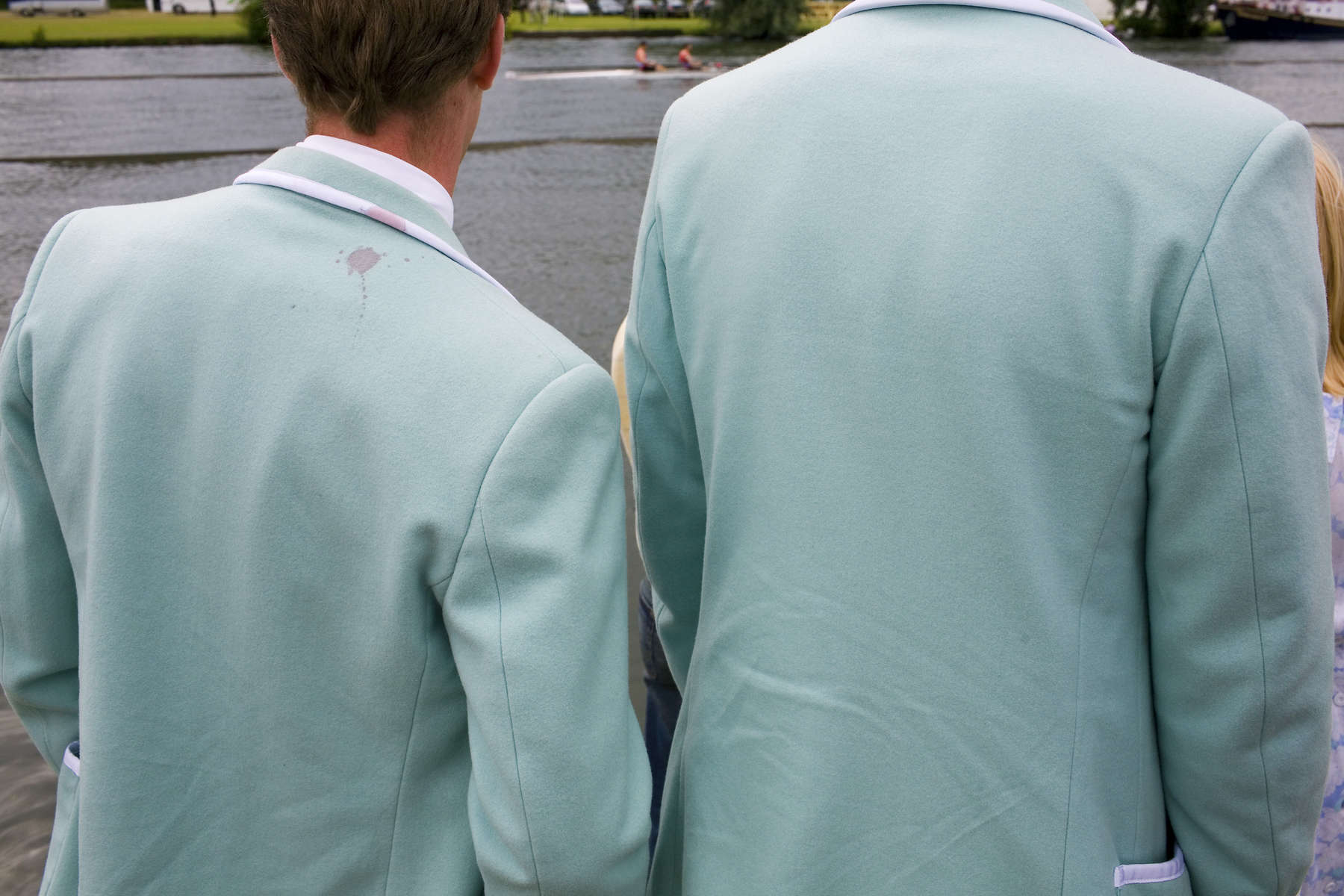 Two boys wearing blazers in the colours of their rowing club watch the racing on the Thames at the Henley Royal Regatta. One of the jackets has what looks like a splash of red wine staining the back. The Regatta is one of the premier events of the English summer season held every year on the north side of the River Thames in south Oxfordshire, England, about 10 miles downstream and north-east from Reading.