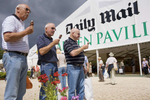Three men eat ice creams as they wait with a bag of plants for their partners outside the Daily Mail sponsored marquee at the Hampton Court Palace annual Royal Horticultural Society summer flower show. Hampton Court Palace is a former royal palace in the London Borough of Richmond Upon Thames. Thomas Wolsey, then Archbishop of York and Chief Minister to the King took over the lease in 1514 and rebuilt the 14th Century manor house over the next 7 years (1515-1521) to form the nucleus of the present palace before Henry VIII took it as his residence.
