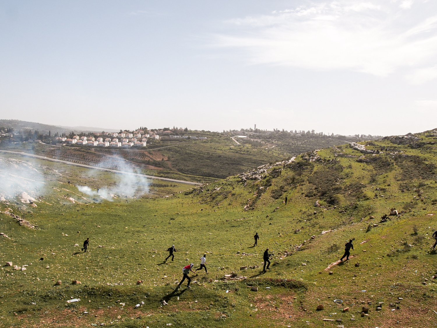 West Bank.  Nabi Saleh.  2013.  Protestors run away as Israeli soldiers fire tear gas at them.  The Israeli settlement of Halimish is in the background. Residents of Nabi Saleh have been protesting the Israeli Occupation every Friday after midday prayers since 2009.  The protests started after Israelis from the nearby settlement of Halimish took over a small spring that had been on Palestinian land.  After widening and adding a bench to the spring, the settlers refused to allow the Palestinians to continue using it.  The weekly protests quickly devolve into rock throwing by the Palestinians and tear gas, rubber bullets, and 'skunk' (a spray mixed with water that has a horrific stench that can linger for weeks) in response by the Israelis.