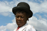 It is a head and shoulders photograph of a 71-year old Afro-Bolivian woman wearing a bowler hat, against the backdrop of a blue sky.