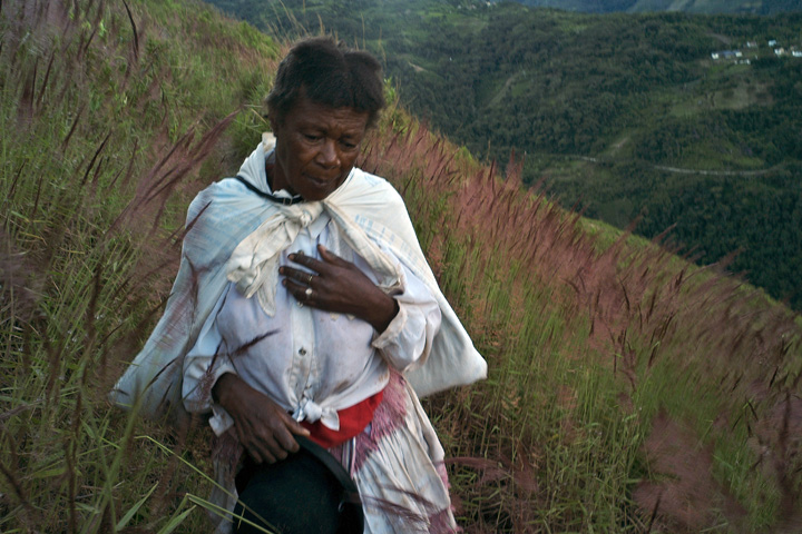 An older Afro-Bolivian woman, holding her bowler hat and with a white sack of coca leaves tied around her shoulders, walks through a lush field on a hill.
