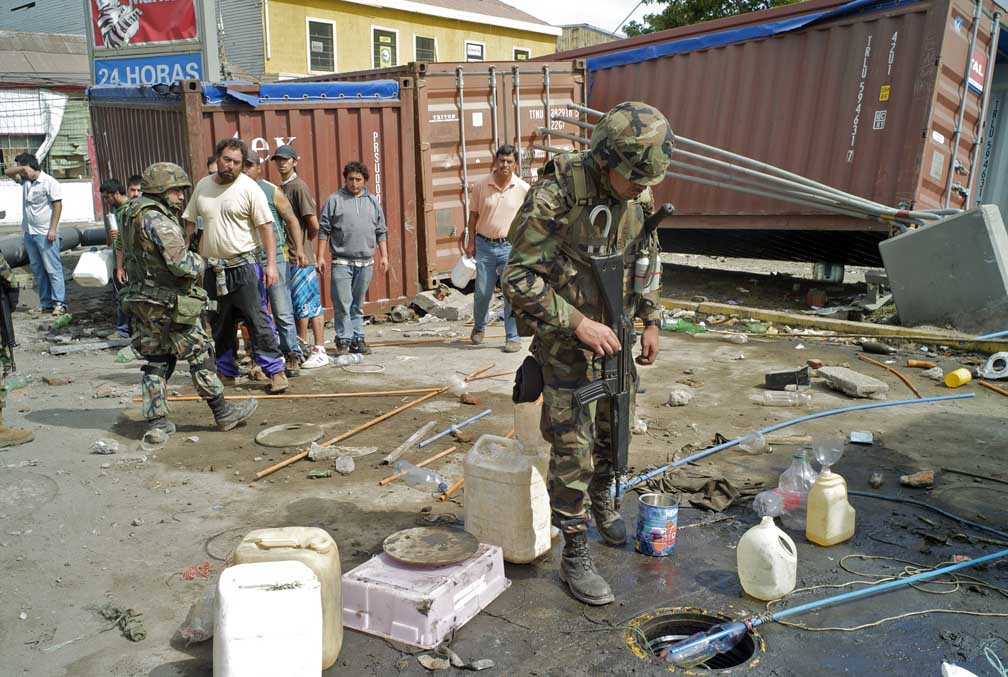 At a gas station a standing armed soldier looks down an underground gas tank, while another soldier detains several men accused of siphoning gas.