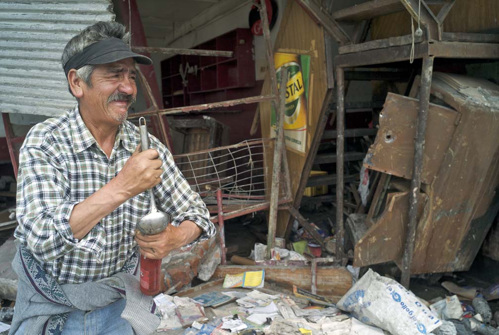 An old man prys open a soda bottle he retrieved from a badly damaged store (visible just behind him).