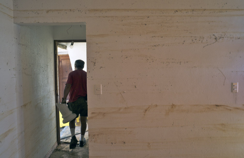 A man holding a broken bathroom fixture is seen walking inside his damaged house; in the foreground a water line reveals that the water reached near the ceiling.