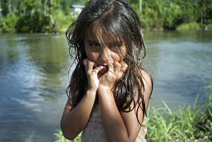 A wet ten-year old girl holds her hands to the lower part of her face and stares into the camera, shortly after exiting a river which is in the background.