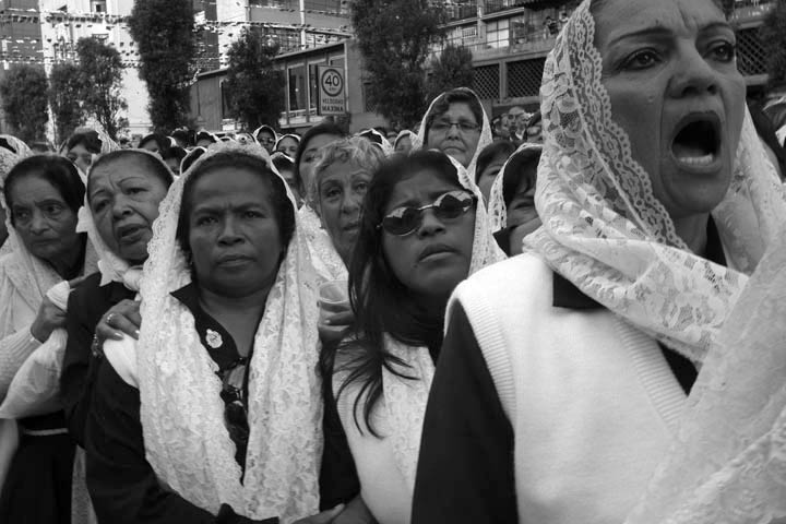 Women dressed in traditional capes watch and sing as a procession carrying a painting adorned with the image of Our Lord of Miracles arrives. 