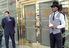 An Afro-Peruvian man dressed in a top-hat, shirt, tie and vest, stands in the doorway of a casino with a white-Peruvian supervisor observing.