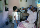 An elderly Afro-Bolivian woman is sitting in a chair in the kitchen while talking to her 7-year old grandson who is standing.